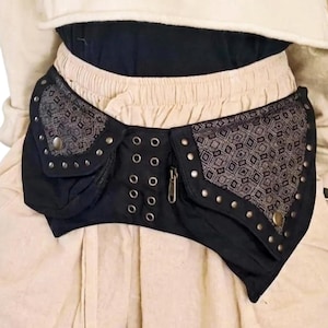Peut inclure: Une ceinture utilitaire noire dotée de deux grandes poches à rabats à motifs géométriques, de clous métalliques argentés et d'un panneau central avec deux rangées verticales d'œillets métalliques. La ceinture inclut une fermeture éclair.