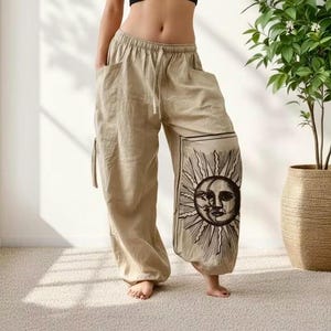 May include: Beige loose-fitting pants with an elastic waistband and a drawstring. The left leg features a large, rectangular black and white graphic print of a sun and moon face with radiating rays. The pants have side pockets and cuffed hems.