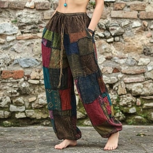 Peut inclure: Pantalon ample présentant un motif patchwork composé de divers panneaux de tissu rectangulaires dans des tons de marron, bleu, vert et rouge. Le pantalon possède une taille élastique avec un long cordon de serrage marron et des poches latérales.
