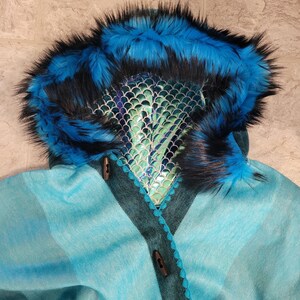 May include: A teal and blue hooded cape with a faux fur trim in blue and black. The hood features a scale-like design and is accented with wooden toggle closures. The cape has a plaid pattern.