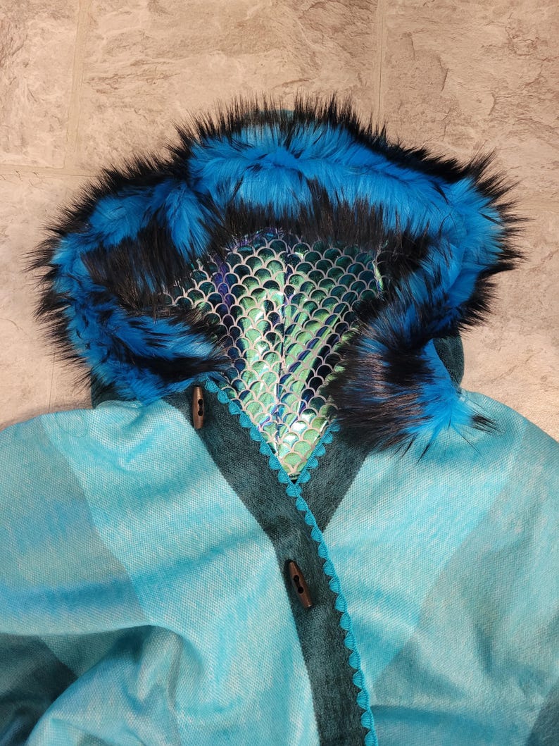 May include: A teal and blue hooded cape with a faux fur trim in blue and black. The hood features a scale-like design and is accented with wooden toggle closures. The cape has a plaid pattern.