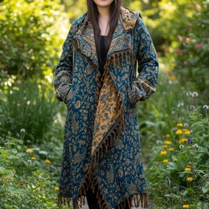 May include: A long-sleeved, open-front coat featuring a teal and mustard yellow paisley pattern. The garment includes a wide collar, a hood, and a hemline finished with decorative fringe detailing. The coat is worn over a black top and black leggings.