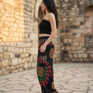 Puede incluir: Una persona con un top corto negro y pantalones negros estampados con un dise&ntilde;o floral colorido. Los pantalones tienen bolsillos y se combinan con chanclas blancas. El fondo presenta una pared de piedra.