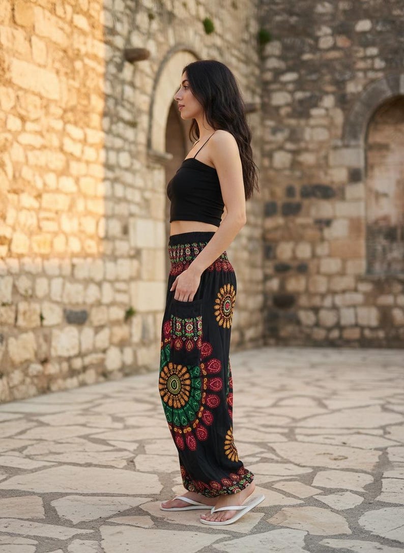Puede incluir: Una persona con un top corto negro y pantalones negros estampados con un dise&ntilde;o floral colorido. Los pantalones tienen bolsillos y se combinan con chanclas blancas. El fondo presenta una pared de piedra.
