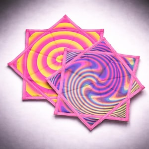May include: Three star-shaped fabric items with vibrant, swirling patterns in shades of pink, yellow, and purple. Each item has a pink border. The designs feature concentric circles and wavy lines, creating a psychedelic effect. The items are arranged on a light background.