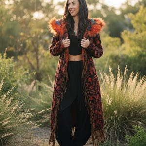 May include: A long, open-front coat featuring a dark fabric with a red floral pattern, long fringe trim along the edges, and a hood lined with vibrant orange faux fur. The garment is worn over a black crop top and black loose-fitting trousers.