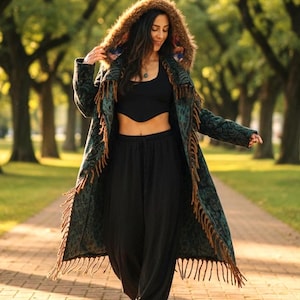May include: A long, patterned coat with a fringed hem and a fur-lined hood. The coat is teal and brown, worn with black pants and a black top. The person is walking on a brick path in a park.