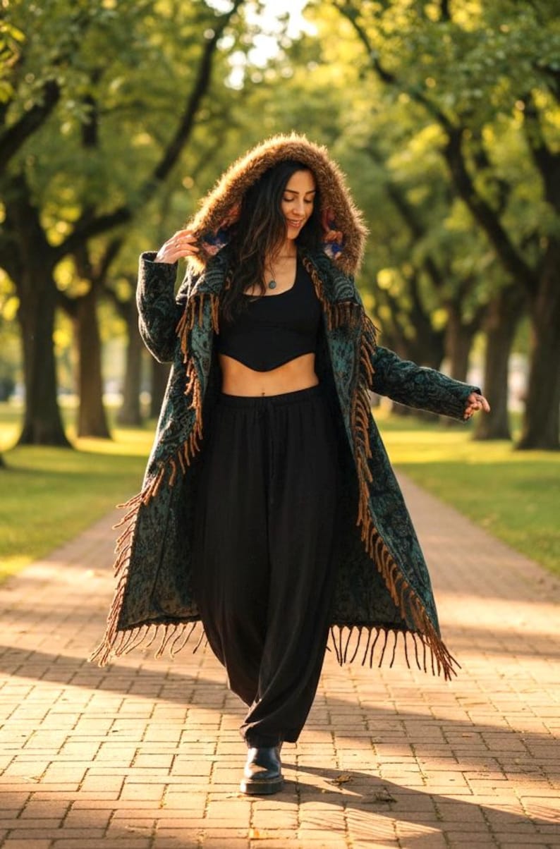 May include: A long, patterned coat with a fringed hem and a fur-lined hood. The coat is teal and brown, worn with black pants and a black top. The person is walking on a brick path in a park.