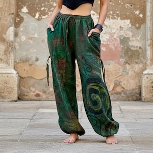 May include: Loose-fitting trousers in a deep forest green hue featuring a dark, swirling spiral pattern on the lower leg. These pants include an elasticized waistband with a drawstring, side pockets, and adjustable ties on the outer seams.