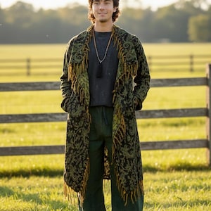 May include: A long, open-front coat featuring a black and olive green paisley pattern with fringe trim along the lapels and hem. The garment is worn over a dark grey t-shirt and loose-fitting dark green trousers, paired with a long beaded necklace.