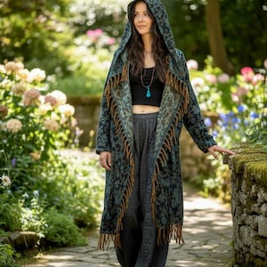 May include: A woman wearing a long, hooded coat with a teal and black floral pattern and fringed edges. The coat is paired with gray pants and a black top. The scene is set in a garden with stone pathways and blooming flowers.