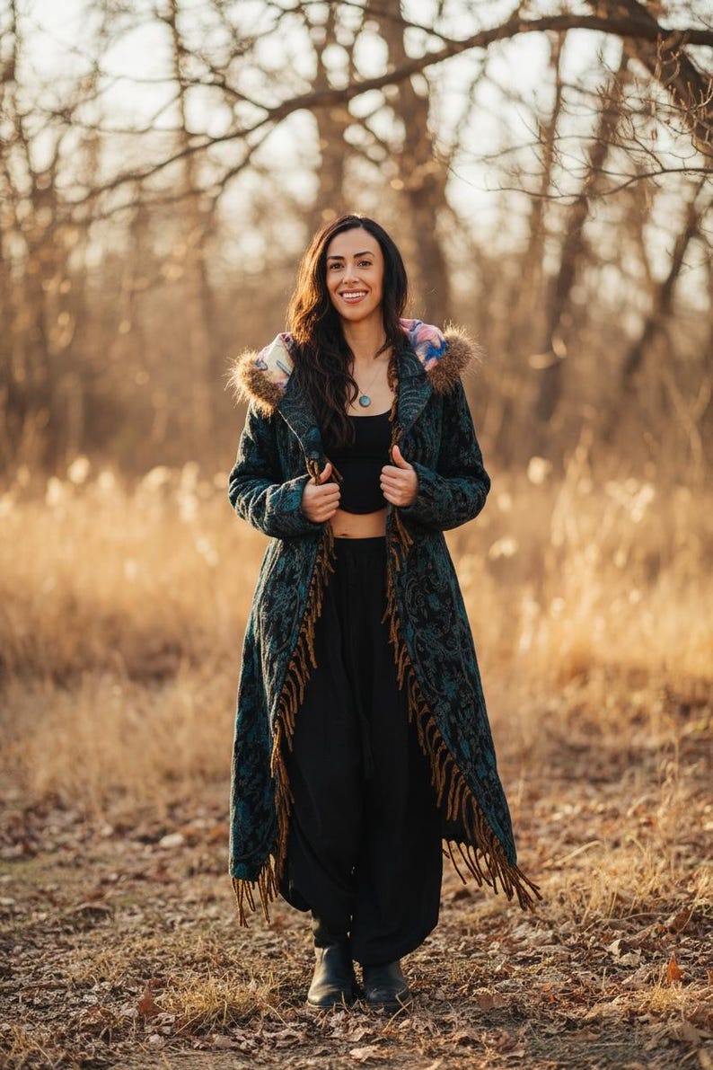 May include: A long, teal coat with a floral pattern and brown fringe. The coat has a fur-lined hood with a colorful design. The person is wearing black pants and a black top. The background is a field with trees.