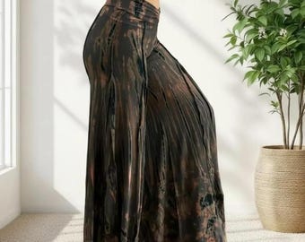 May include: Wide-leg palazzo pants with a tie-dye pattern in shades of brown, black, and a hint of peach. The high-waisted pants are made of a flowing fabric, creating a relaxed and comfortable fit. The pants are shown in a well-lit room with a potted plant.