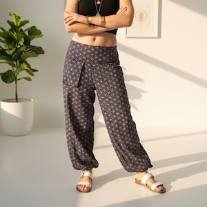 Op de afbeelding: Een persoon draagt een zwarte haltertop en een broek met patroon met een geometrisch ontwerp. De broek is donker met een herhalend wit patroon. Witte sandalen met bruine zolen maken de outfit compleet. Gouden sieraden zijn ook zichtbaar.