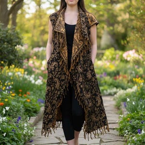 May include: A long, sleeveless black and gold patterned vest with fringe trim. The vest has an intricate paisley design and is worn over a black top and leggings. The person is walking on a stone path in a garden.