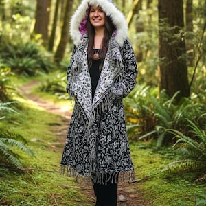 May include: A long black and white patterned coat with a hood lined with white faux fur and a pink interior. The coat has a shawl collar and fringe trim. The person is standing on a path in a forest.