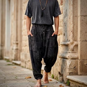 May include: A charcoal gray short-sleeved t-shirt with a distressed finish paired with black drop-crotch trousers featuring a subtle marbled pattern, large side pockets, and an elasticized waistband with a drawstring, worn with a long beaded necklace.