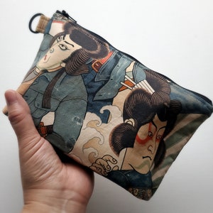 May include: A zippered pouch with a Japanese-inspired design featuring two figures in blue and white clothing against a beige background with waves. The pouch has a black zipper and a black loop.