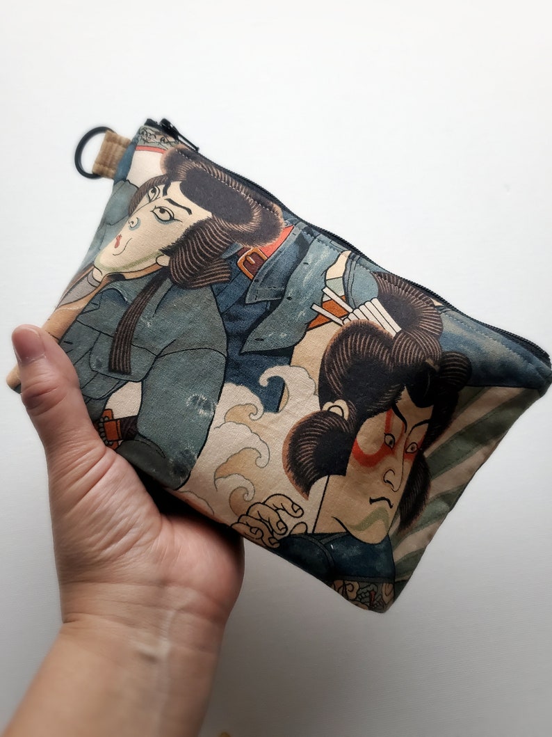 May include: A zippered pouch with a Japanese-inspired design featuring two figures in blue and white clothing against a beige background with waves. The pouch has a black zipper and a black loop.