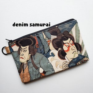 May include: A zippered pouch with a colourful Japanese-inspired print featuring two samurai warriors in blue and white clothing. The pouch has a black zipper and a black ring attached to the zipper pull.