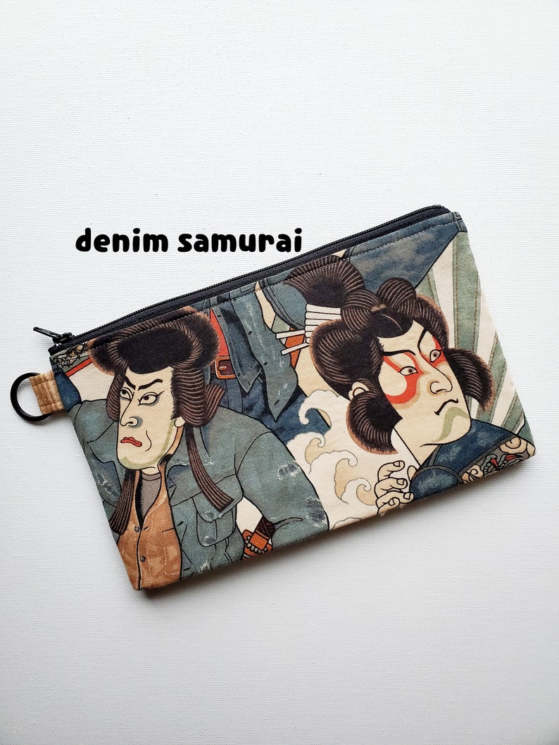 May include: A zippered pouch with a colourful Japanese-inspired print featuring two samurai warriors in blue and white clothing. The pouch has a black zipper and a black ring attached to the zipper pull.