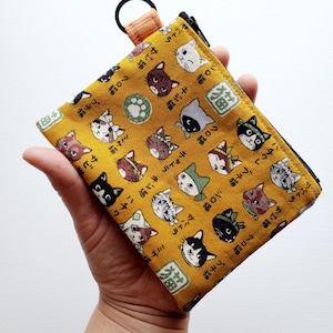 May include: Yellow cat-themed wallet with a black zipper and a small orange loop. The wallet features a repeating pattern of various cat faces and Japanese text. The cats are in various colors, including black, brown, and white.