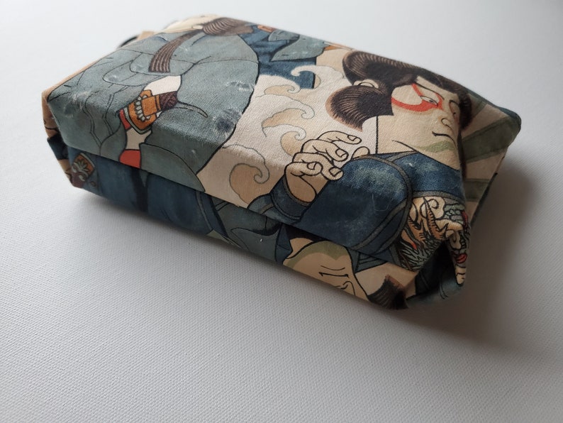 May include: A small, rectangular pouch with a Japanese-inspired print featuring figures in blue and beige clothing. The pouch is made of fabric and has a zipper closure.
