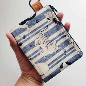 May include: A rectangular fabric pouch featuring a pattern of cats on a background of blue and white horizontal stripes. The pouch includes a black zipper closure along the top edge and a small orange fabric loop with a black metal ring attached.