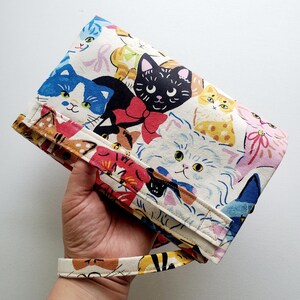 May include: A white canvas clutch bag with a colorful print of cats. The bag has a long strap and a flap closure.
