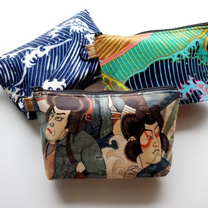 May include: Three zippered pouches with different designs. The first pouch is blue with a white wave pattern. The second pouch is green and yellow with a wave pattern. The third pouch is brown with a Japanese-style illustration of two people.
