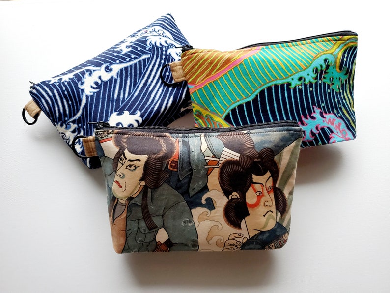 May include: Three zippered pouches with different designs. The first pouch is blue with a white wave pattern. The second pouch is green and yellow with a wave pattern. The third pouch is brown with a Japanese-style illustration of two people.