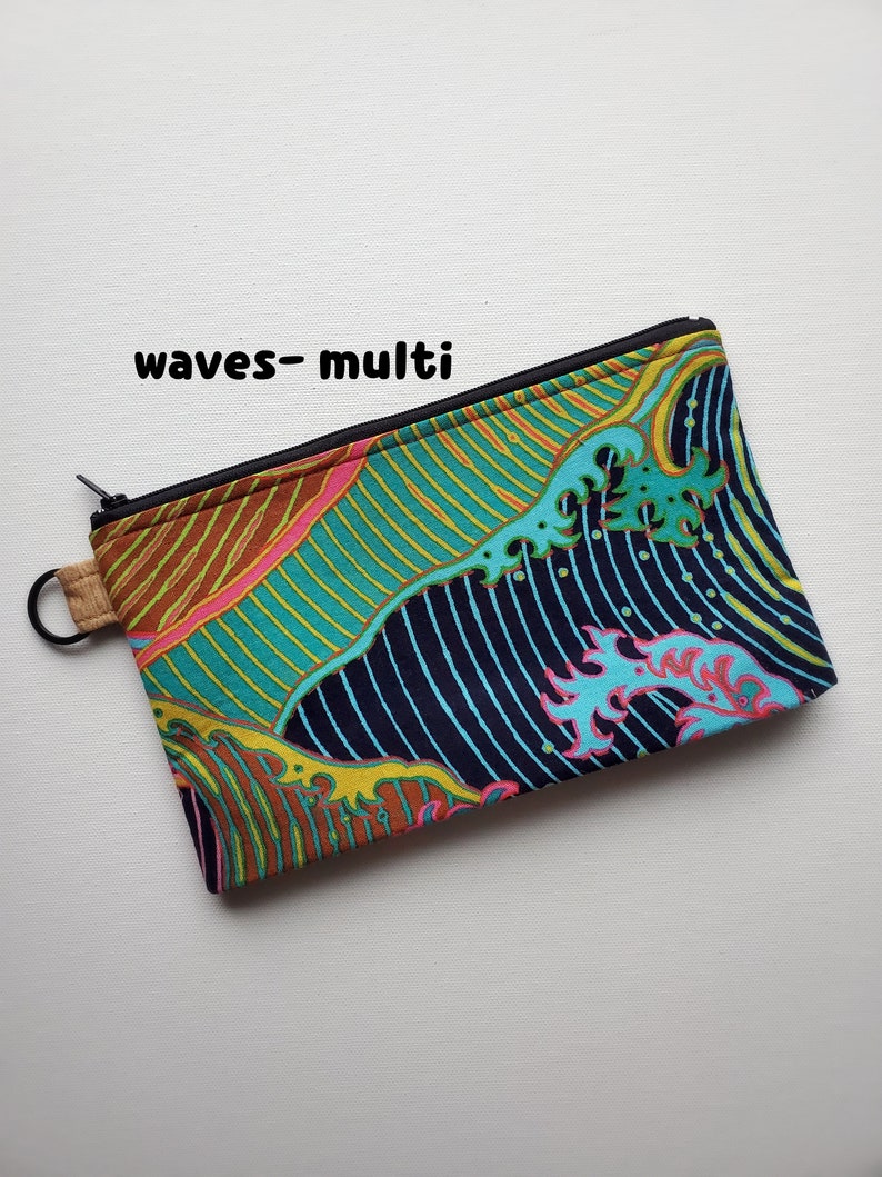 May include: A zippered pouch with a colourful wave pattern in shades of blue, green, yellow, pink, and brown. The pouch has a black zipper and a brown strap.