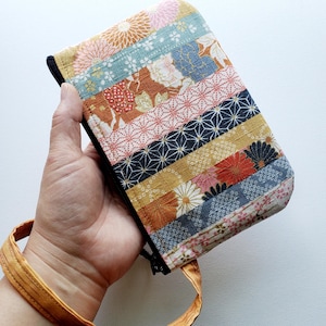 May include: A colorful, rectangular fabric pouch with a black zipper and a matching wrist strap. The pouch is made of various patterned fabric strips in shades of pink, blue, gold, and brown. The wrist strap is a light orange color.