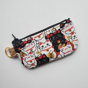 May include: A small white zippered pouch with a black zipper and a gold key ring. The pouch is covered in a pattern of black and white lucky cat figures with red accents.