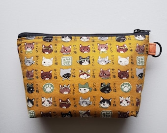 May include: A yellow zippered pouch with a black zipper and a black and orange pull tab. The pouch is covered in a pattern of cats with Japanese text. The text includes the word "猫" which means "cat" in Japanese.