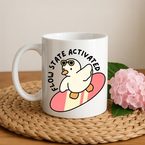 May include: White ceramic mug featuring a cartoon duck wearing sunglasses and riding a surfboard. The mug has the text "FLOW STATE ACTIVATED" in a circular design. The mug is on a woven mat.