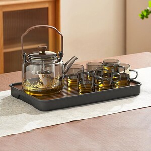 May include: A glass tea set with a teapot and six cups on a black tray. The teapot has a wooden handle and a gold fish design. The cups are clear with a handle and contain a golden liquid.