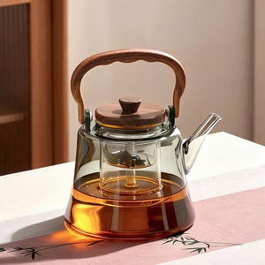 Può includere: Teiera in vetro trasparente con manico e coperchio in legno. La teiera ha un infusore interno ed è riempita di liquido color ambra. Il manico e il coperchio sono in legno scuro. Progettata per l'infusione del tè.