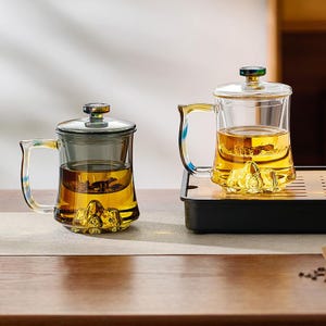 Può includere: Due infusori per tè in vetro trasparente con manici e coperchi. Gli infusori sono riempiti di tè color ambra e presentano un design a forma di montagna alla base. Uno è su un vassoio nero, l'altro su una superficie neutra.