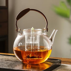 May include: A clear glass teapot with a brown wooden handle and a built-in infuser. The teapot is filled with amber-colored tea. The handle is arched and attached to the pot with metal brackets. The teapot sits on a dark wooden tray.