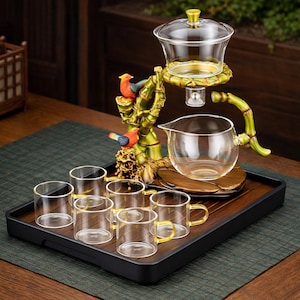 May include: A clear glass tea set with a bamboo-style stand, a teapot, and six small cups. The stand features decorative birds and a wooden base. The set is presented on a black tray, perfect for serving tea.