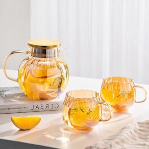Könnte beinhalten: Ein Teegeschirr aus Glas mit einer Teekanne und zwei Tassen. Die Teekanne hat einen goldenen Deckel und Griff, und die Tassen haben Griffe. Das Set ist mit einer gelben Flüssigkeit und Orangenscheiben gefüllt. Das Set steht auf einer weißen Oberfläche.