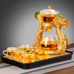 Puede incluir: Un juego de té con forma de dragón dorado con una tetera de vidrio, tazas y una bandeja. El juego de té incluye una tetera de vidrio, una jarra de vidrio y seis tazas pequeñas de vidrio, todas llenas de té. El cuerpo del dragón está intrincadamente detallado.