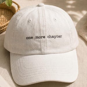 May include: A white baseball cap featuring the embroidered black text "one more chapter" centred on the front panel. The cap has a structured crown, a curved brim, and a button at the top, constructed from a solid-coloured fabric material.