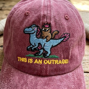 May include: A maroon baseball cap featuring an embroidered graphic of a blue dinosaur with a small brown cat wearing a pink crown on its back. Below the graphic, the text THIS IS AN OUTRAGE! is embroidered in yellow capital letters.