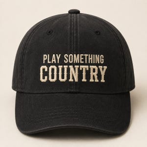 May include: A black baseball cap featuring the embroidered text PLAY SOMETHING COUNTRY in a light beige, serif font. The cap has a structured crown, a curved brim, and a button at the top centre, showcasing a classic six-panel design.