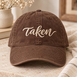 May include: A brown baseball cap with a distressed finish featuring the word taken embroidered in a light beige cursive script across the front panel. The cap has a curved brim, stitched eyelets, and a structured crown design.