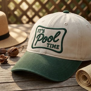 May include: A beige and green baseball cap with the text "It's Pool Time" embroidered on the front. The cap has a green brim and a green button on top. Other hats and natural elements are in the background.