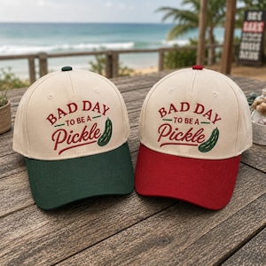 May include: Two baseball caps with the embroidered text "BAD DAY to be a Pickle". One cap has a green peak, the other a red peak. Both caps have a cream crown and a green pickle graphic.