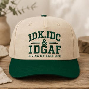 May include: A beige baseball cap with a dark green brim and dark green embroidered text on the front that reads IDK, IDC & IDGAF LIVING MY BEST LIFE. The cap features a structured crown with a top button and stitched eyelets for ventilation.
